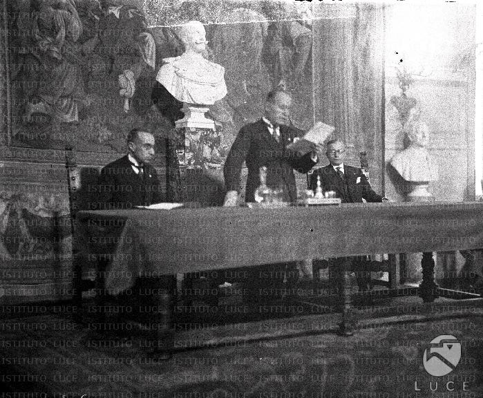 Benito Mussolini legge un discorso nella sala di un palazzo. Accanto a ...