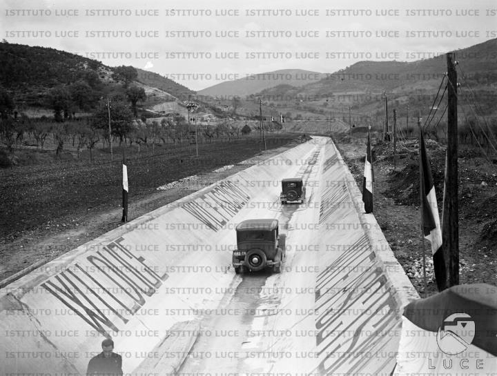 Due automobili, l'una con Mussolini a bordo, transitano lungo il canale ...