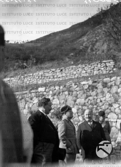Mussolini ripreso tra due personalità lungo una strada, ai piedi di un ...