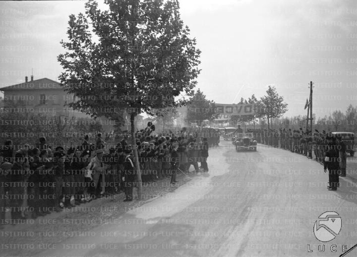 Mussolini, in piedi a bordo di una automobile, percorre una strada di ...