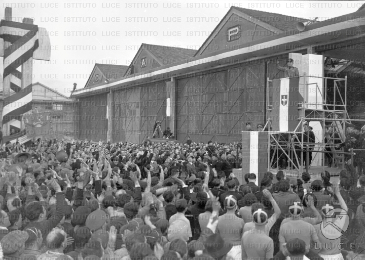 Mussolini in piedi su un palco fa il saluto fascista e viene acclamato ...