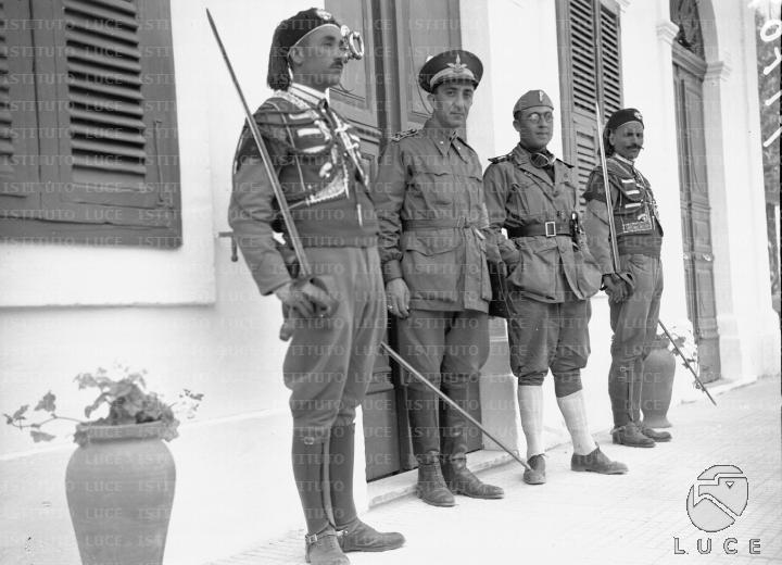 Un ufficiale della milizia e un autiere del Regio esercito ritratti  all'esterno di un edificio coloniale - forse una caserma - presidiato da  due sciumbasci degli zaptiè in grande uniforme - Archivio