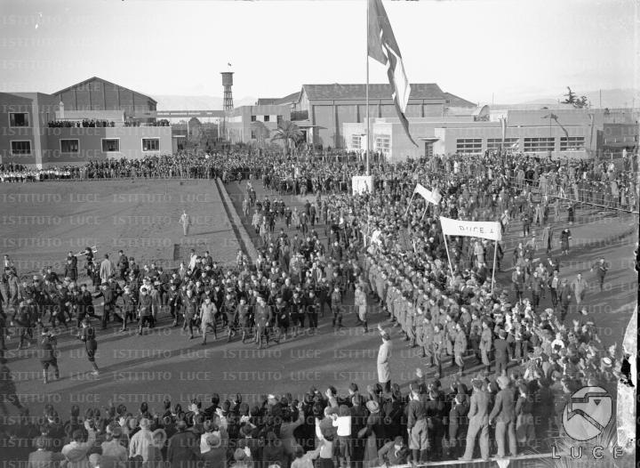 Mussolini, accompagnato da un seguito di gerarchi, sfila lungo un viale ...