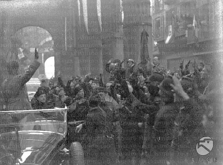 Mussolini, in piedi a bordo di un'automobile, fa il saluto fascista ...