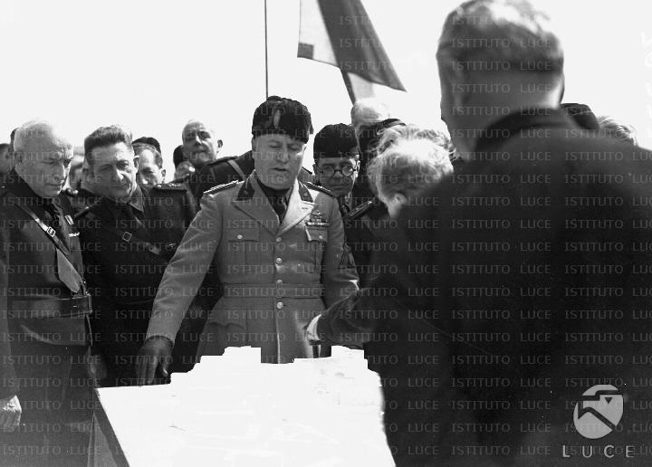 Mussolini, circondato all'aperto da un gruppo di autorità in orbace ...